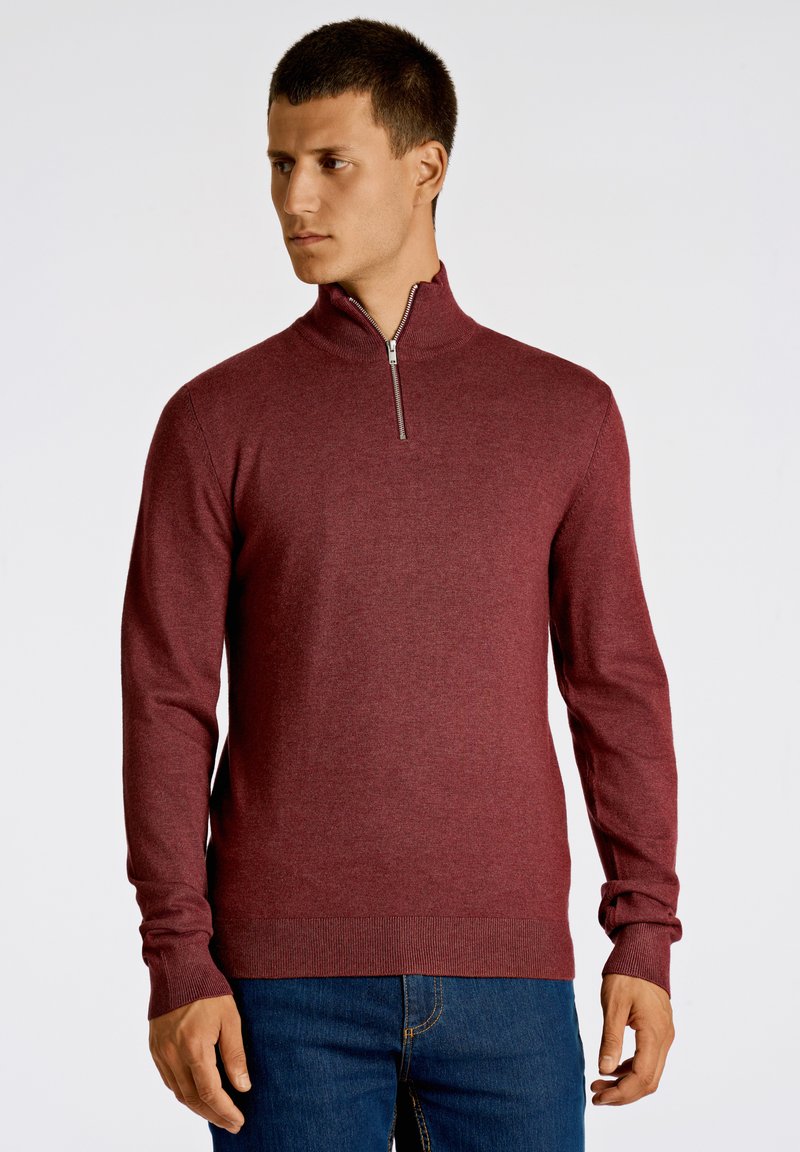 Maglione con zip in borgogna, realizzato in morbido tessuto a maglia. Caratterizzato da maniche lunghe e polsini a costine, abbinato a jeans in denim blu.