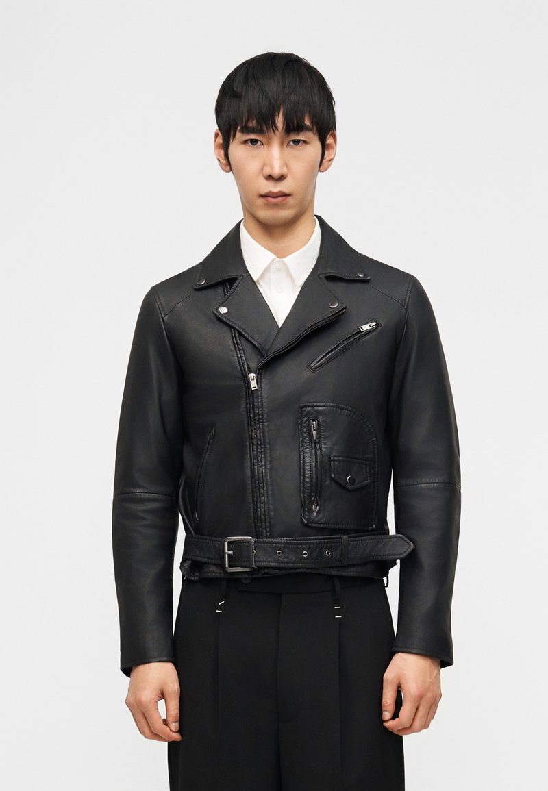 Άνδρας που φοράει μαύρο δερμάτινο μπουφάν μοτοσικλετιστή με φερμουάρ στις τσέπες, λεπτομέρεια με ζώνη και λευκό πουκάμισο με γιακά από κάτω, στέκεται μπροστά σε απλό φόντο.