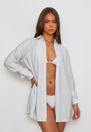 Camicia argentata e lucida con bottoni, caratterizzata da maniche lunghe, una vestibilità morbida e un profondo scollo a V, indossata sopra un completo bikini coordinato.