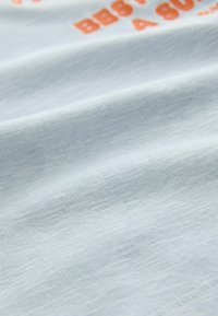 Lyseblåt stof i nærbillede, der viser en blød tekstur med delvist synlig orange tekst i store bogstaver.