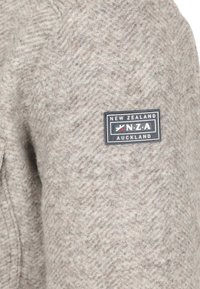 Haringbone-patroon, lichtbruine wol sweater met een zwart label met de tekst "NEW ZEALAND N.Z.A AUCKLAND" op de linkerschouder.