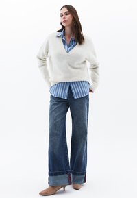 Maglione bianco a maglia, camicia azzurra a righe con bottoni, jeans in denim a gamba larga con orli risvoltati e décolleté marroni a punta. Sfondo semplice.