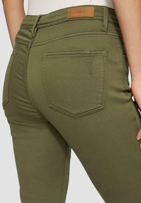 Olive krāsas šauras džinsi ar gludu faktūru, ar ādas birku jostas vietā un diviem aizmugurējiem kabatām ar izšūtu detalizāciju.