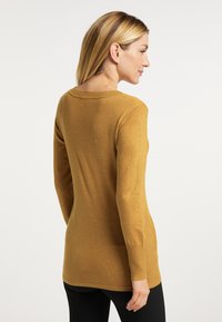 Mosterdkleurige lange mouwen trui met een ronde halslijn, geribbelde manchetten en een aansluitende pasvorm, gedragen over zwarte leggings.