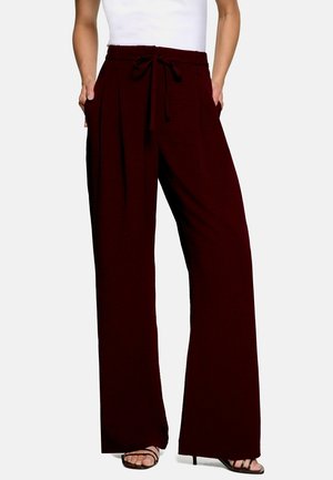 Femme portant un pantalon large taille haute bordeaux foncé avec une ceinture à nouer et des talons ouverts noirs, debout les mains dans les poches.