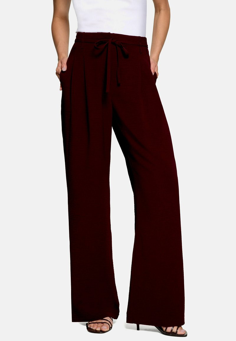 Femme portant un pantalon large taille haute bordeaux foncé avec une ceinture à nouer et des talons ouverts noirs, debout les mains dans les poches.