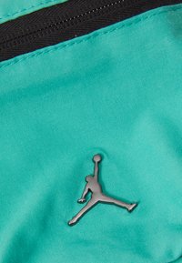 Tessuto turchese con una superficie liscia, dotato di una cerniera nera e un accento del logo Jumpman in argento. Focalizzazione su colori vivaci e design elegante.