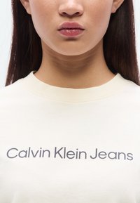 Ανοιχτό μπεζ μπλουζάκι με στρογγυλή λαιμόκοψη, με το κείμενο "Calvin Klein Jeans" τυπωμένο σε σκούρο γκρι. Ομαλή υφή υφάσματος, μινιμαλιστικός σχεδιασμός.