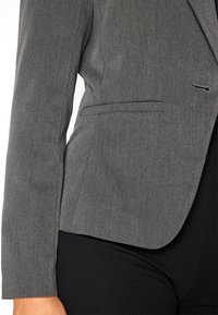 Šedý blazer s texturovanou tkaninou, který se vyznačuje elegantním designem, přiléhavým střihem, detailem na jedním knoflíku a postranními kapsami. Černé kalhoty jsou viditelné dole.