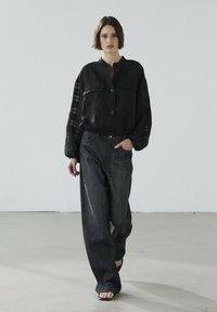 Zwarte lange mouwen button-up shirt van textuurstof met twee borstzakken, gecombineerd met donkere denim wijde pantalons en open teen sandalen.