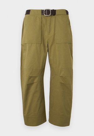 Ung kvinde står med hænderne i lommerne iført hvid kortærmet crop top, olivengrønne bukser med vide ben og sort bælte samt brune loafers.