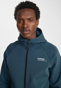 Felpa blu scuro con zip, realizzata in tessuto liscio e con colletto alto. Presenta maniche raglan e un piccolo logo bianco sul petto.