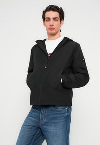 Μαύρο μπουφάν με φερμουάρ και κουκούλα, ελαφρύ υλικό, με ελαστικές μανσέτες και πλαϊνές τσέπες. Φορεμένο πάνω από ένα λευκό πουκάμισο και μπλε τζιν.