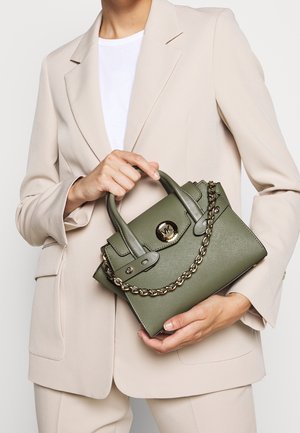 Personne portant un costume beige tenant un sac à main vert olive avec une chaîne dorée et un fermoir logo métallique rond.