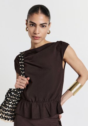 Haut marron texturé avec un ourlet peplum et un col asymétrique. Le mannequin tient un sac tiss é noir et blanc et porte un bracelet manchette en or.