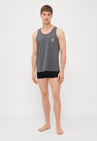 Pilkas medvilnės tank topas su apvaliu iškirpimu, kuriame yra mažas logotipo akcentas ant krūtinės. Modelis derina jį su juodais šortais, stovėdamas basomis.