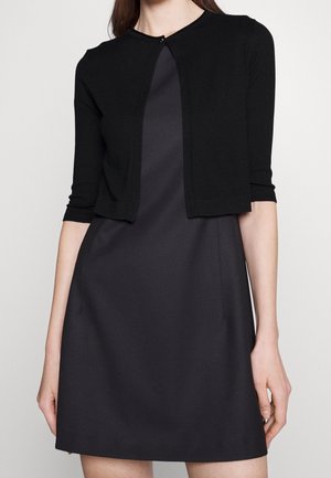 Nainen, joka pukeutunut mustaan lyhyeen neuletakkiin, jossa on kolme neljänneksen mittaiset hihat, mustan hihattoman mekon päällä vaaleaa yksiväristä taustaa vasten.