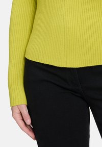 Rippstrick-Pullover in leuchtendem Limettengrün mit langen Ärmeln und enganliegender Silhouette, kombiniert mit schwarzen strukturierten Hosen.