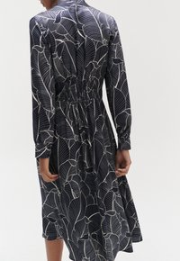 Dunkles Blumenkleid mit gerafftem Taillenbereich, langen Ärmeln und fließendem Design. Hergestellt aus leichtem Stoff mit einem Blättermuster in Weiß.