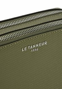 Portafoglio in verde oliva con texture e doppie cerniere argentate. Presenta il logo in rilievo "LE TANNEUR 1898" sulla superficie. Materiale resistente.