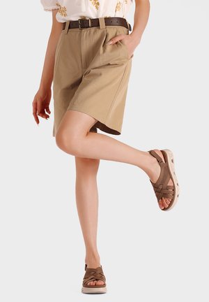 Donna che indossa pantaloncini beige al ginocchio con cintura, camicia bianca con ricamo giallo e sandali marroni con punta aperta, sollevando leggermente una gamba.