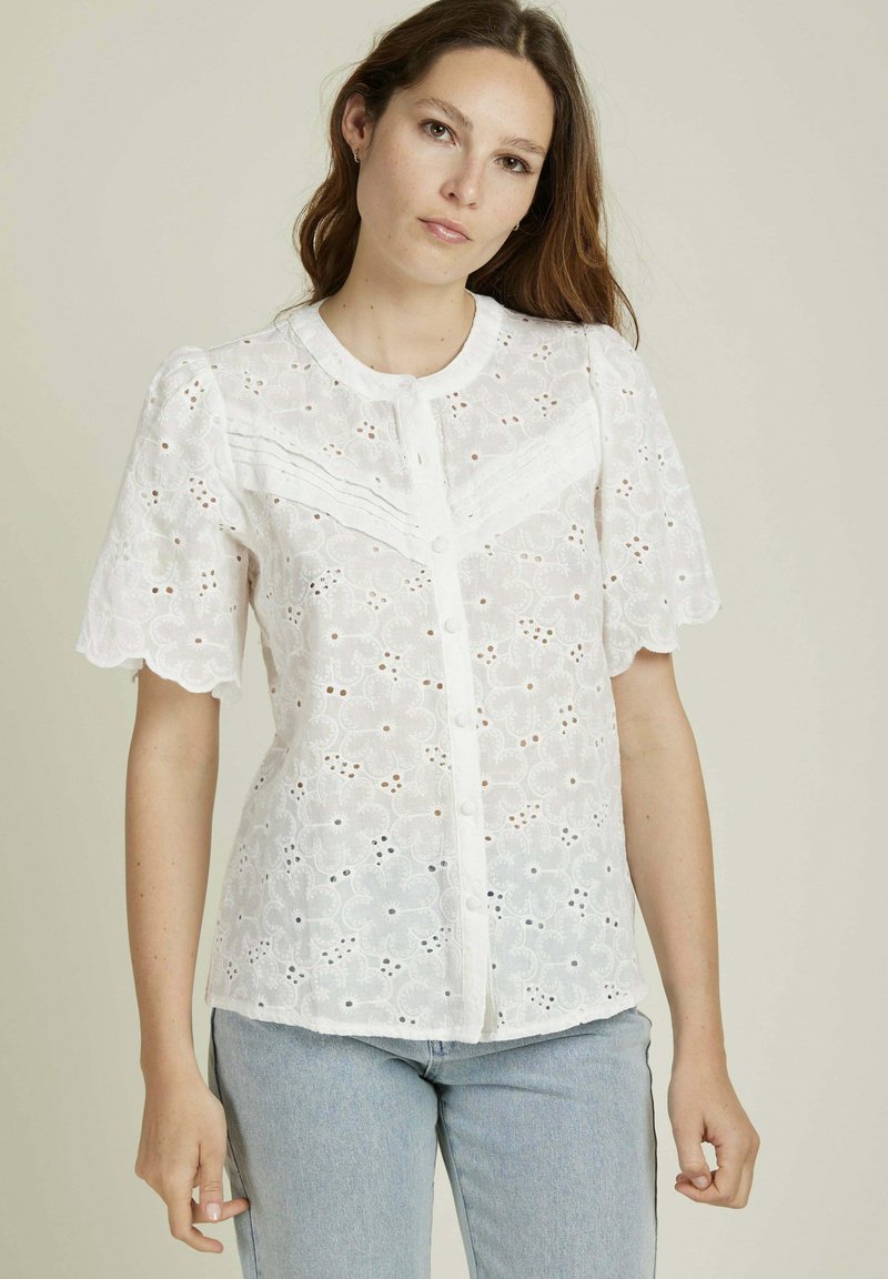 Blouse blanche en broderie anglaise, avec des manches courtes bouffantes, des bords festonnés, un devant à boutons et des motifs floraux découpés.