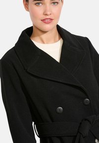 Czarny dwurzędowy płaszcz z szerokim kołnierzem, z paskiem do wiązania i czarnymi guzikami. Wykonany z teksturowanej tkaniny.