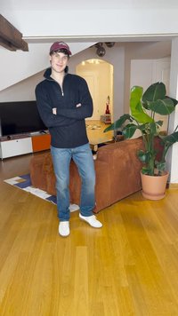 Sweat à zip noir, jeans bleus et baskets blanches. Cadre intérieur avec un canapé en velours côtelé marron et une plante en pot à ses côtés.