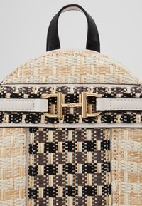 Sac à dos tissé beige et noir de forme ronde. Il est doté d'une fermeture à boucle dorée, d'un matériau texturé et de motifs contrastants.