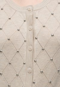 Cardigan beige en tricot avec un motif losange diagonal, petites clous ronds argentés aux intersections et quatre boutons beiges au centre.