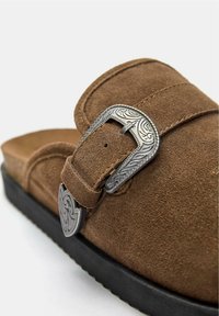 Sandalo scivoloso in suede marrone con una fibbia ornamentale argentata e una suola nera testurizzata. La vista ravvicinata mette in evidenza i dettagli del materiale e del design.