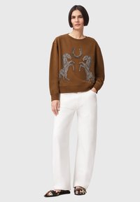 Καφέ φούτερ με κεντημένα σχέδια αλόγων και πέταλο, σε συνδυασμό με λευκό παντελόνι φαρδιάς γραμμής και παπούτσια slip-on με σχέδια.