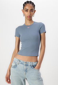 Fitted blue ribbed t-shirt with short sleeves and a round neckline, featuring a small red logo on the chest, paired with light blue jeans.