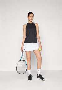 Mustamata ilma varrukat sportlik topp ümmarguse kaelusega, millel on valge tenniseseelik Adidas logo. Hoidis tennisereketit ja palli.