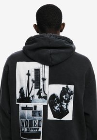 Felpa nera con grandi patch grafiche. Presenta texture, stili fotografici vari e elementi bianchi e scuri a contrasto. Vista posteriore mostrata.