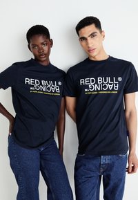 Marineblå t-shirts med korte ærmer, prydet med hvid og gul "RED BULL RACING" tekst. Båret med denimjeans, hvilket præsenterer et afslappet look.