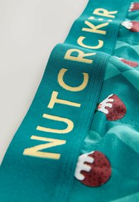 Tyrkis talje bånd med "NUTCRACKER" i gult, prydet med et festligt mønster af brune ornamenter med hvide accenter og røde prikker.