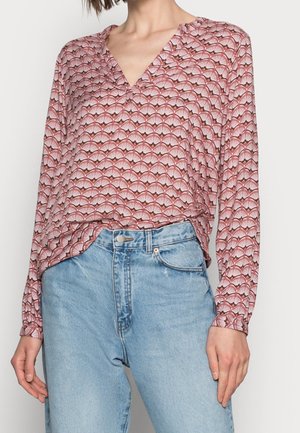 Femme portant un chemisier à manches longues à motifs roses, rentré dans un jean taille haute bleu clair, debout devant un fond blanc uni.