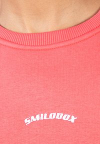 Korallrosa sweatshirt med ribbad rund halsringning, med vit broderad text "SMILODOX" i en böjd form framtill. Mjuk tygtextur.