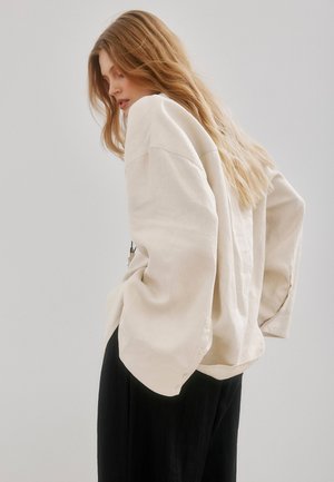 Vrouw met lang blond haar die een oversized crème jas en zwarte broek draagt, staand met handen in de zakken tegen een effen achtergrond.