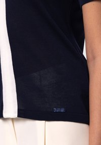 Navyblå stickad topp med ett vitt vertikalt rand, ribbad nederkant och en liten märke nära botten på vänster sida.