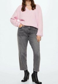 Růžový svetr s výstřihem do V, volným střihem a žebrovanými manžetami, doplněný šedými džíny s vysokým pasem a černými kotníkovými botami na klínu, vytvářející vrstvený vzhled.