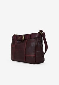 Bolso de piel marrón con bolsillos laterales y una correa desmontable. Presenta un detalle de cinturón en la parte superior con herrajes de latón. Textura suave.
