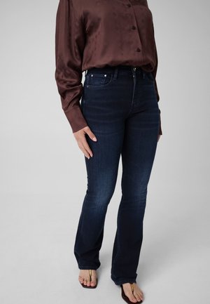 Personne portant un jean taille haute bleu foncé avec une chemise marron à manches longues et des sandales marron à bout ouvert sur un fond uni.