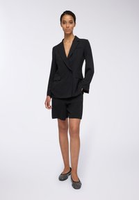 Blazer nero con bottoni doppi e tasche frontali, abbinato a pantaloni corti neri. Il modello indossa ballerine nere decorate. Tessuto dalla texture liscia.