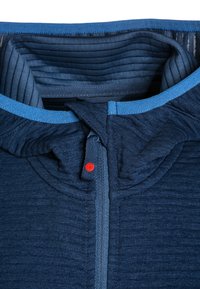 Marinblå tröja med strukturerad tyg, med dragkedja, kontrasterande blå kant och en liten röd detalj vid dragkedjan.