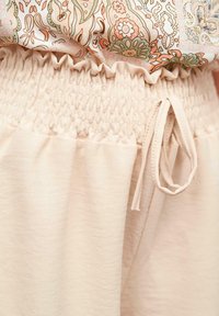 Tissu beige clair avec une texture froncée à la taille, présentant un lien à cordon. Brillance subtile, finition douce et détail de taille volanté.