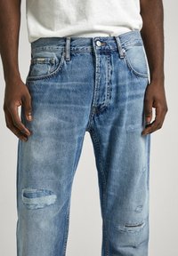Jeans de mezclilla de color azul claro con un corte holgado, que presentan detalles desgastados, múltiples bolsillos y un cierre clásico de botón.
