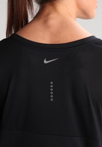 Musta urheilupaita, jossa on teksturoitu kangas, pyöreä kaula-aukko ja heijastava Nike-logo takana. Sisältää viisi pyöreää ilmanvaihtoaukkoa logoa alapuolella.