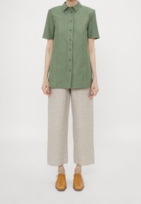 Camicia verde a maniche corte con chiusura a bottoni e cuciture sul petto, abbinata a pantaloni larghi in lino beige e scarpe marroni con lacci.